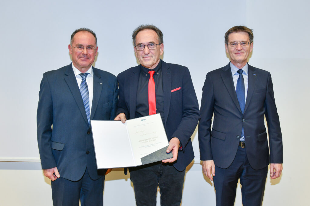 Prejemnik priznanja z rdečo kravato in odprto listino na skupinski fotografiji. Praznovanje 20. obletnice Medicinske fakultete UM.