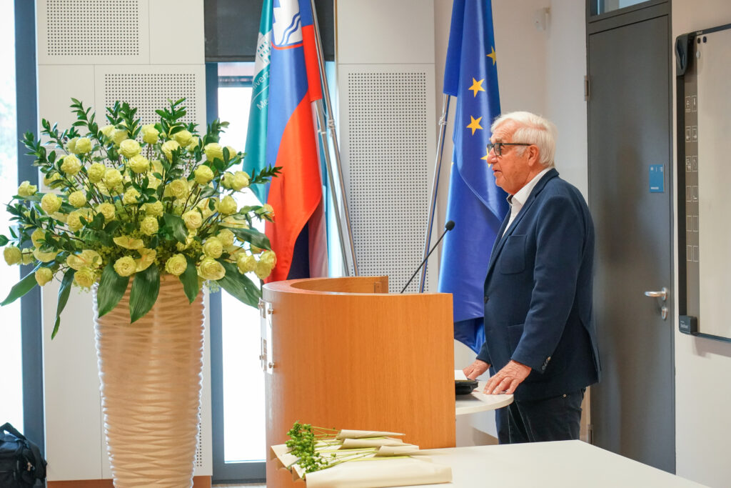 Govorec nagovarja zbrane ob zastavah in cvetličnem aranžmaju. Podelitev diplom Medicinske fakultete UM, 15. junija 2023.